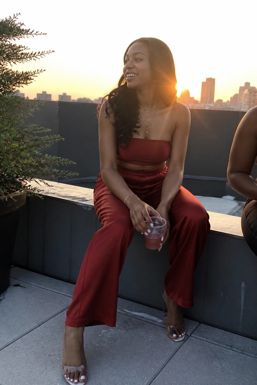 Woman in red two-piece outfit on rooftop ledge at sunset holding drink