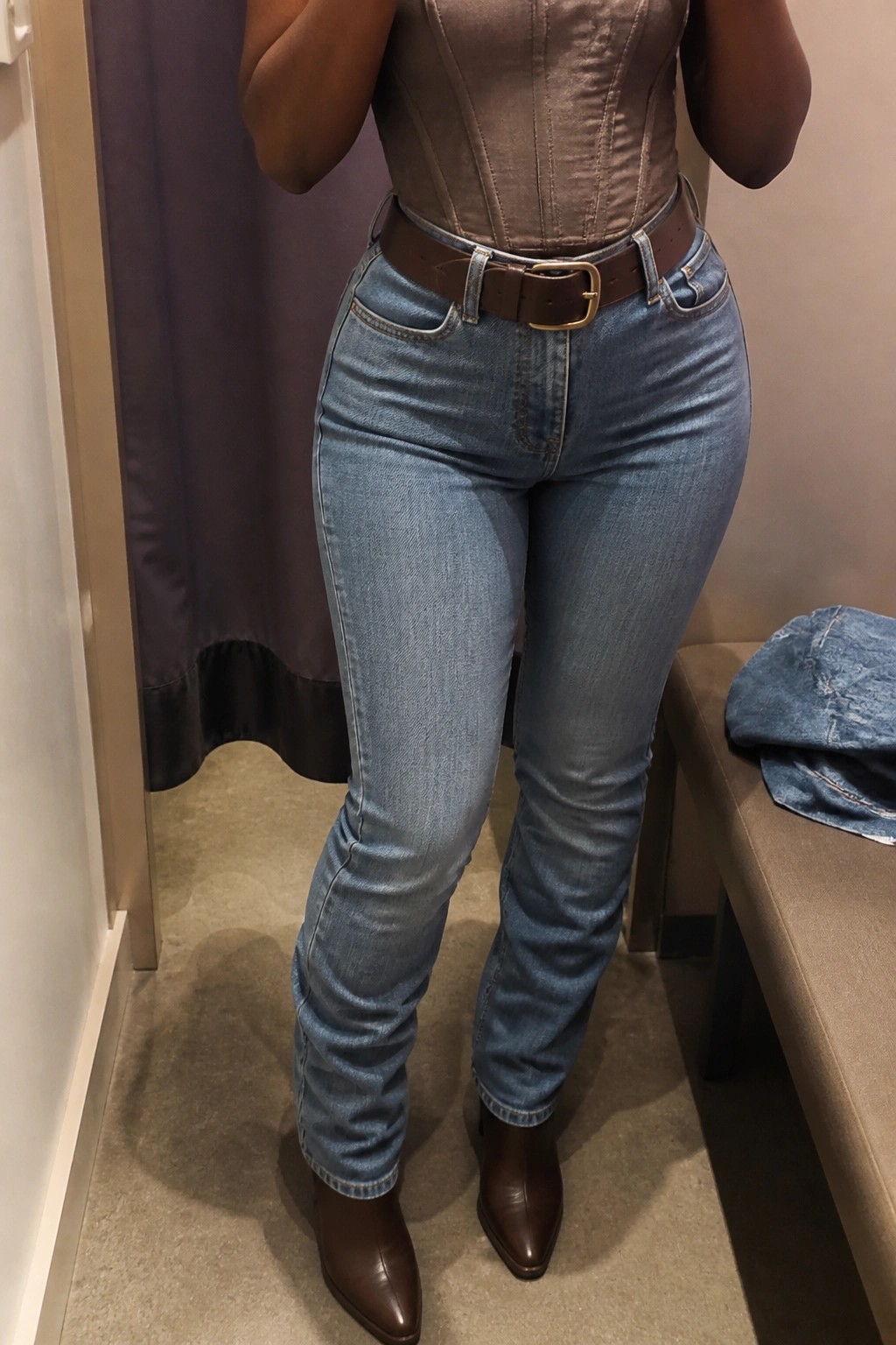Woman in brown corset top, blue jeans, brown belt and boots in fitting room