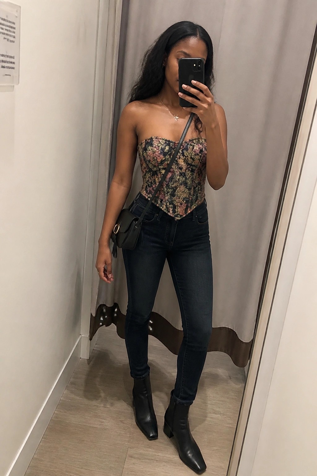 Woman in floral corset top and jeans taking mirror selfie in fitting room