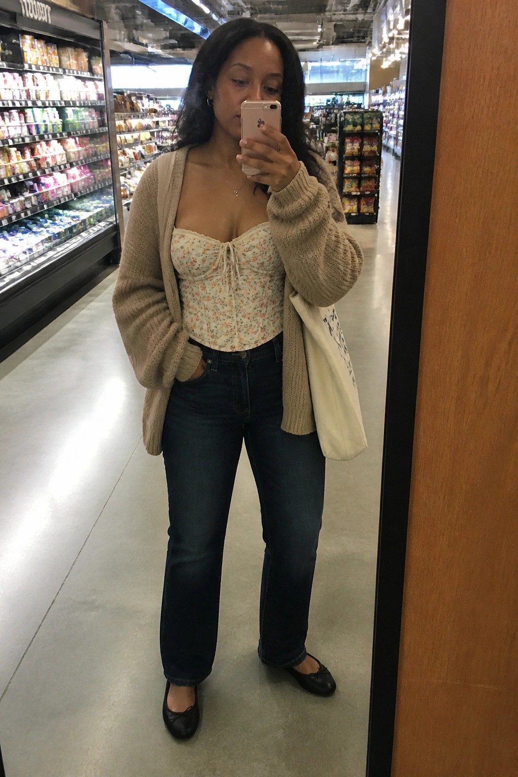 Woman in beige cardigan over floral top takes mirror selfie in grocery store.
