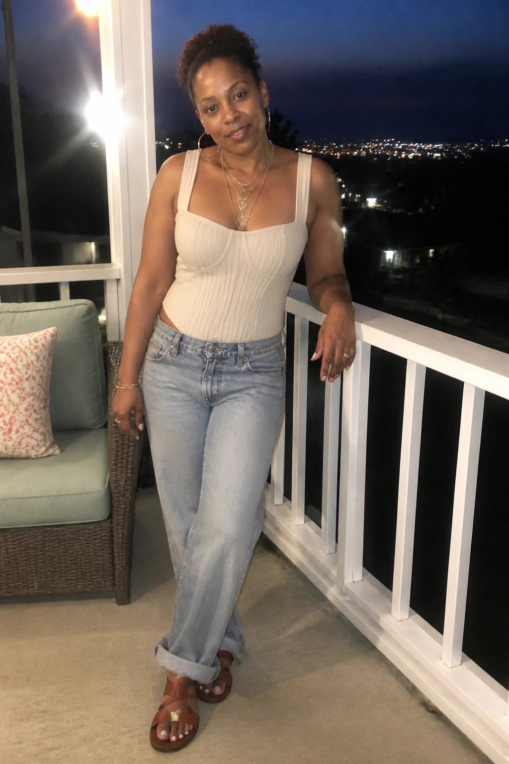 Woman in beige ribbed top and light-wash jeans on balcony overlooking city lights at night