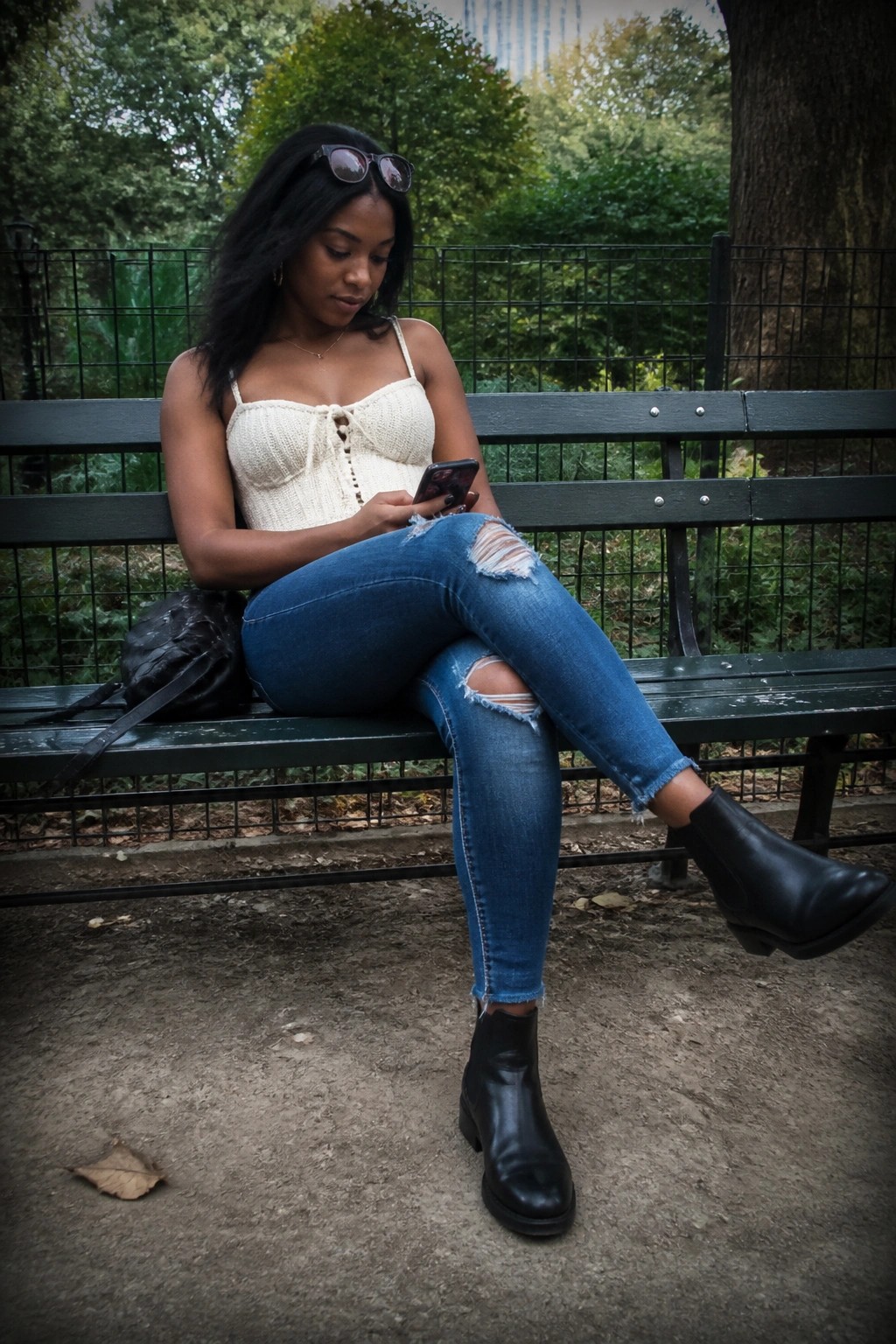 Woman in white knit top and ripped jeans using phone on park bench