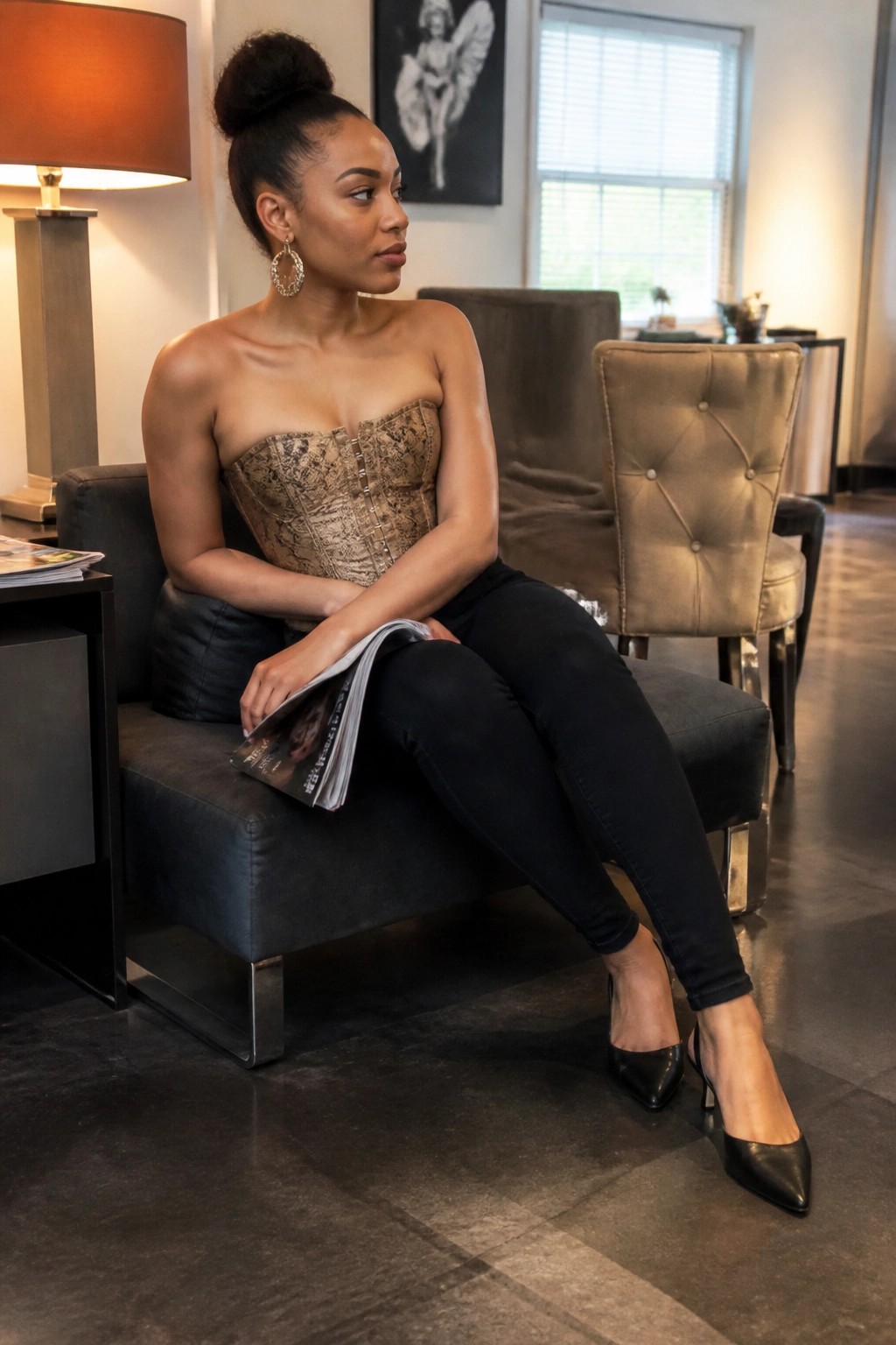 Woman wearing a strapless snakeskin patterned corset top tucked into black skinny jeans with pointed black heels while seated and holding a magazine
