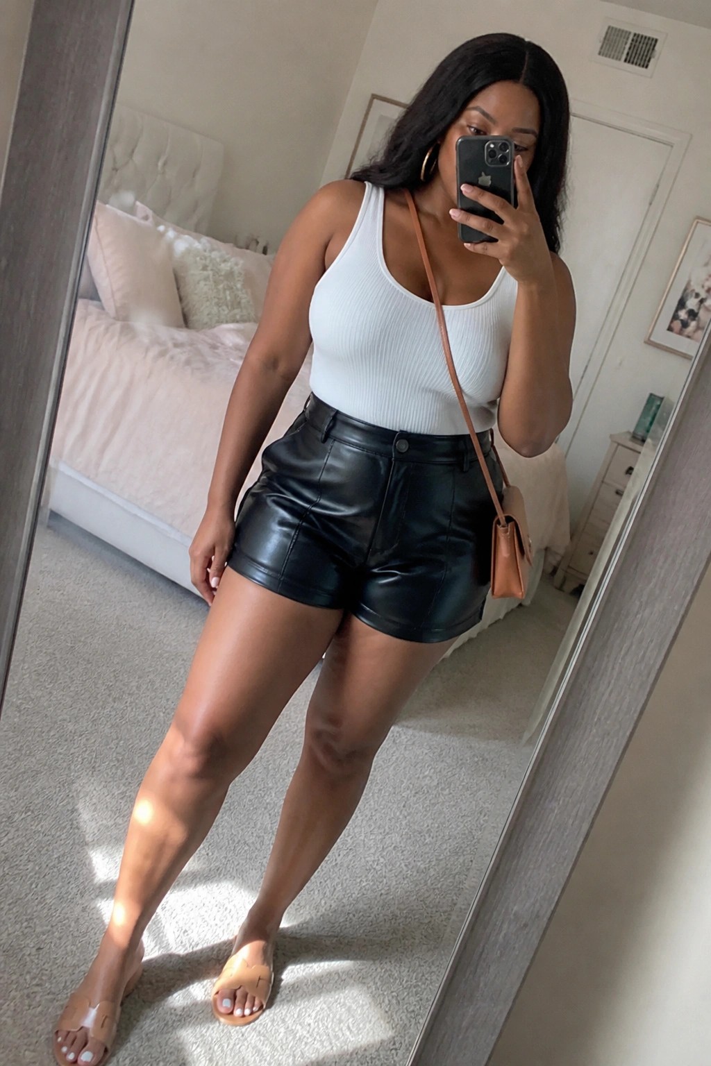 Woman in white tank top and black leather shorts taking mirror selfie in bedroom