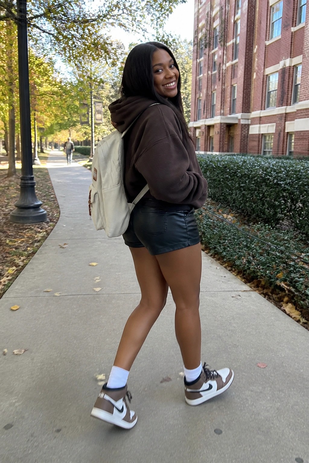 A Black woman wearing an oversized brown hoodie, black leather shorts, white socks, and brown and white high top sneakers with a beige backpack.