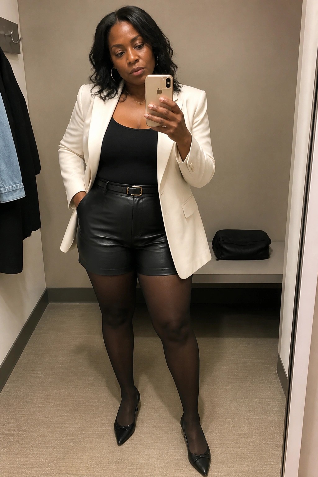 Woman in white blazer and black leather shorts taking mirror selfie in fitting room.