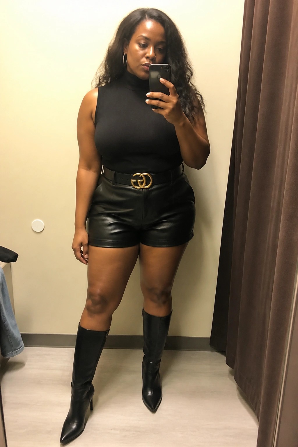 Woman in black turtleneck and leather shorts with gold buckle belt taking mirror selfie