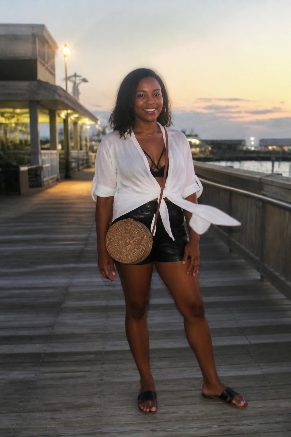 Woman in tied white shirt and black shorts with woven bag on pier at sunset