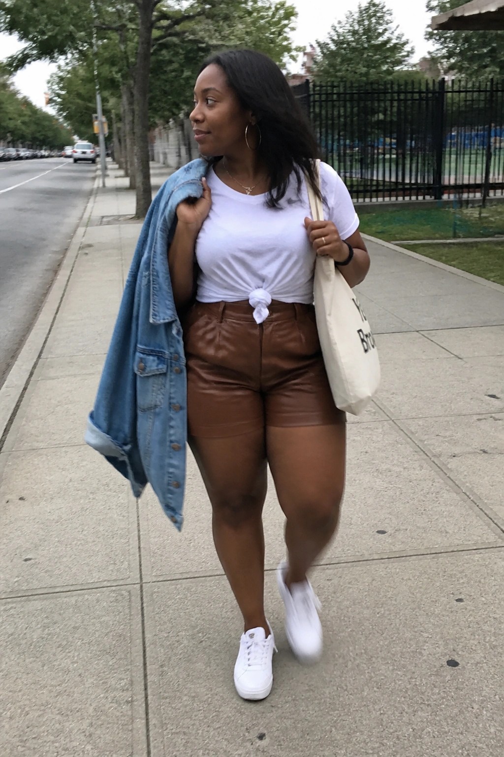 Woman in white tied t-shirt and brown shorts carries denim jacket and tote on sidewalk