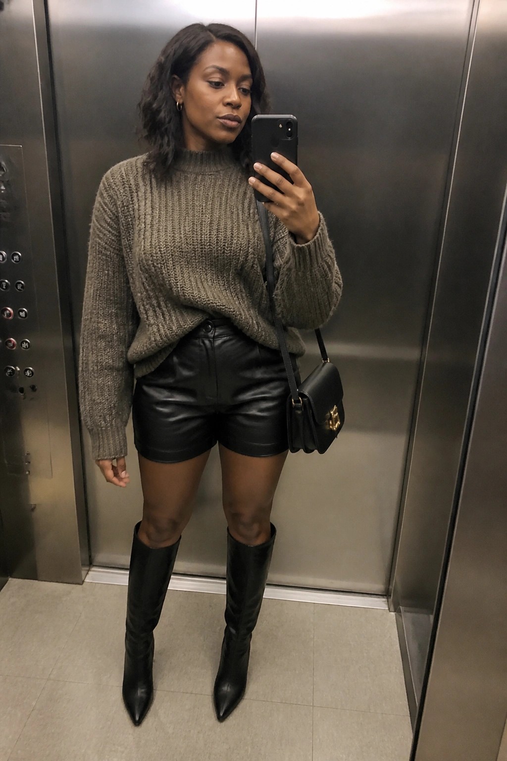 Woman in chunky knit sweater and black leather shorts taking elevator mirror selfie