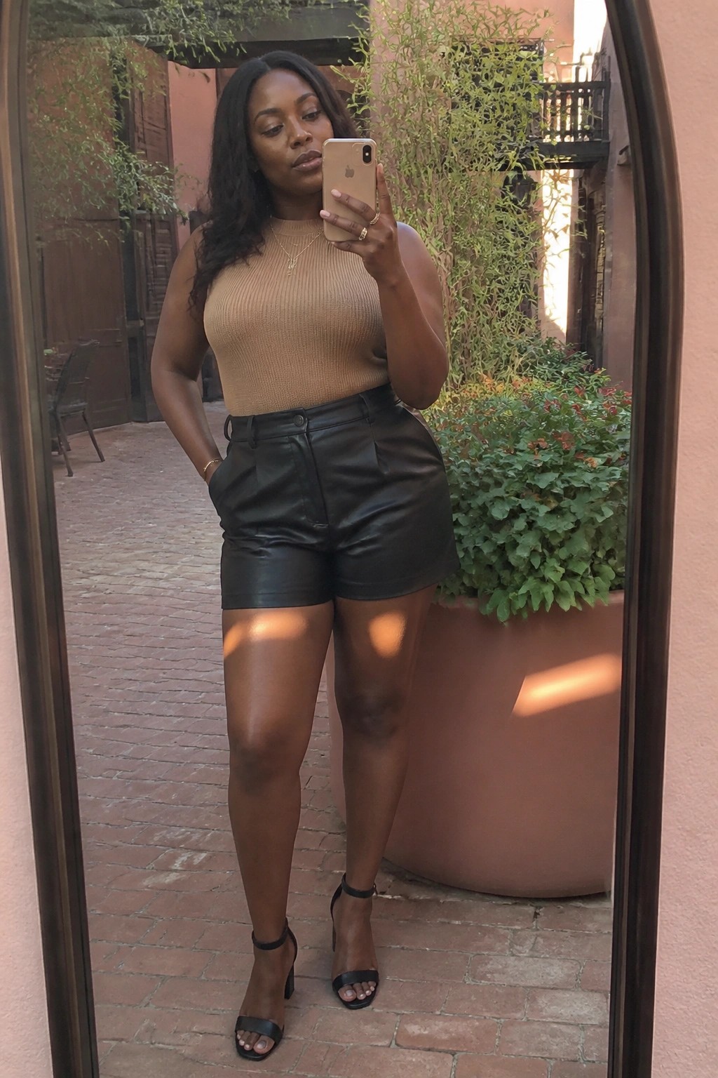 Black woman in mirror selfie wearing high-waisted black leather shorts, beige ribbed sleeveless tank, black block-heel sandals, and layered gold necklaces.