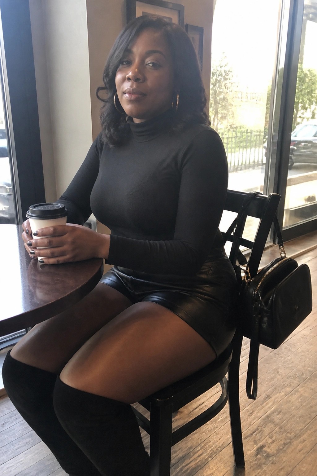 Black woman seated indoors wearing a fitted black turtleneck top tucked into black leather shorts with black thigh high boots and holding a coffee cup next to a black handbag on the chair.