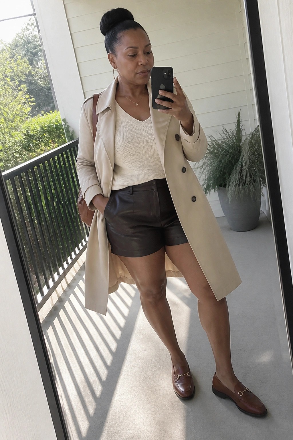 Black woman wearing an open beige trench coat over a cream V-neck knit sweater and dark brown leather shorts, paired with brown loafers featuring gold accents, standing on a porch while holding a phone for a mirror selfie.
