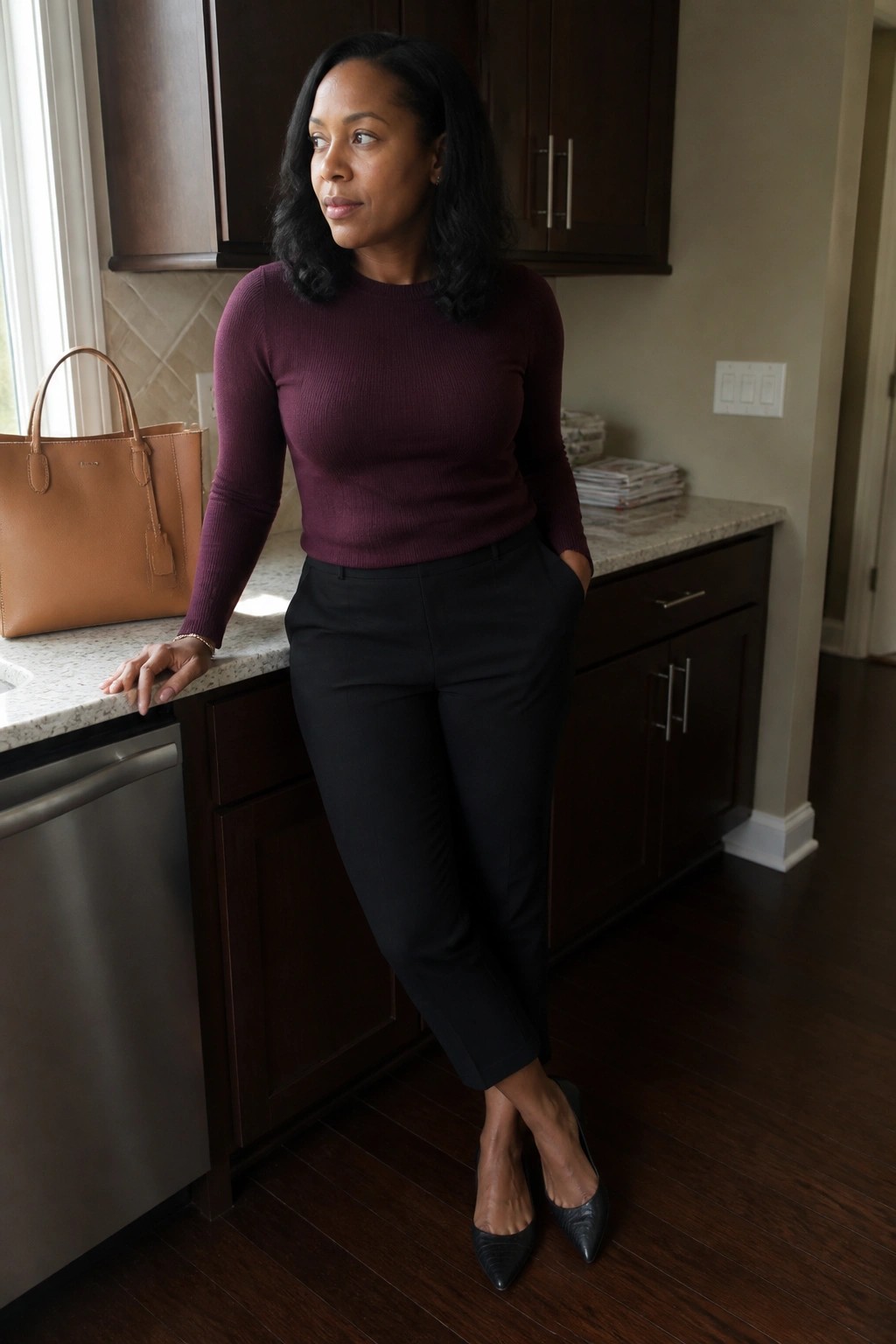 Woman in maroon sweater and black pants leaning on kitchen counter beside brown handbag