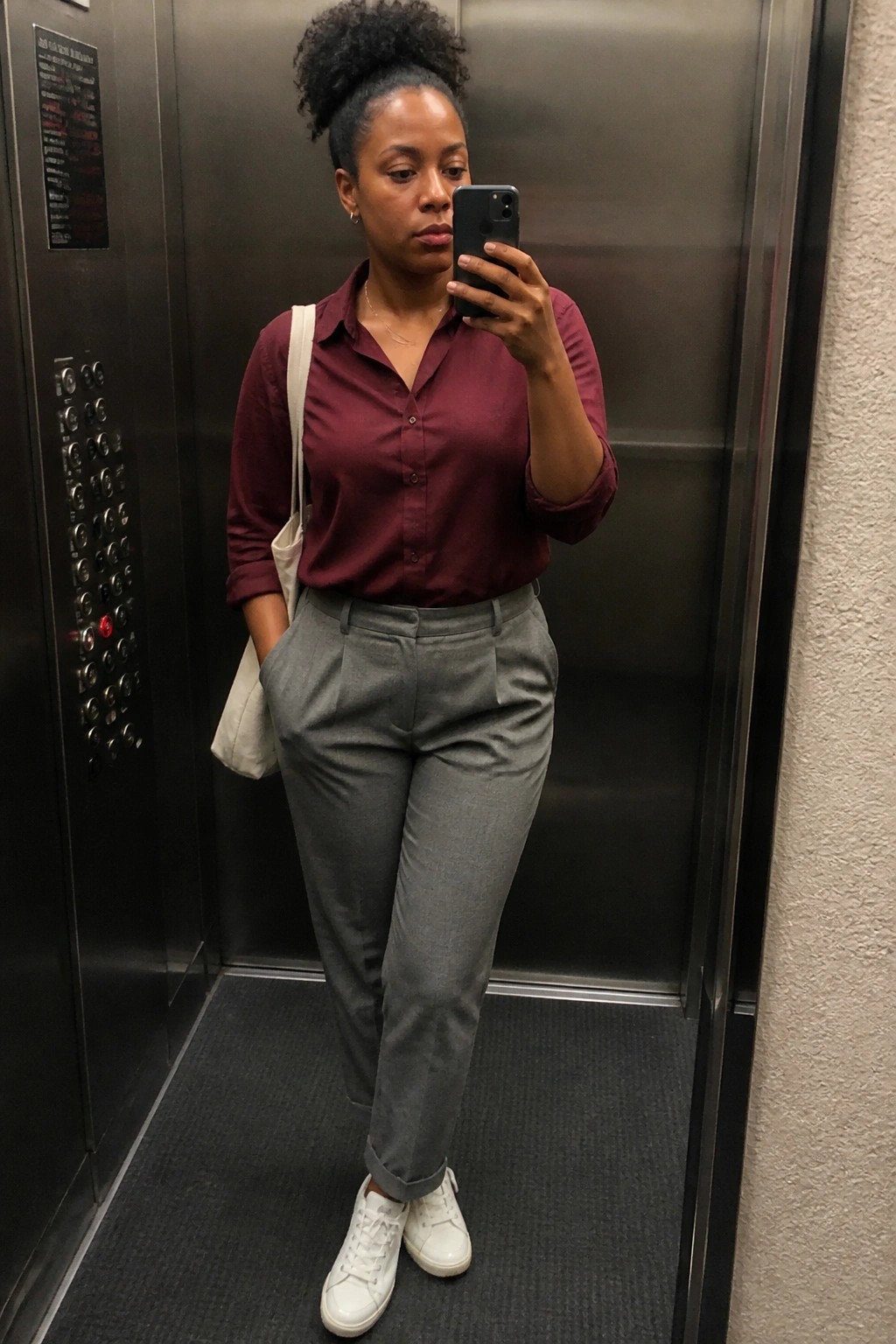 Woman in maroon shirt and gray trousers taking phone selfie in elevator