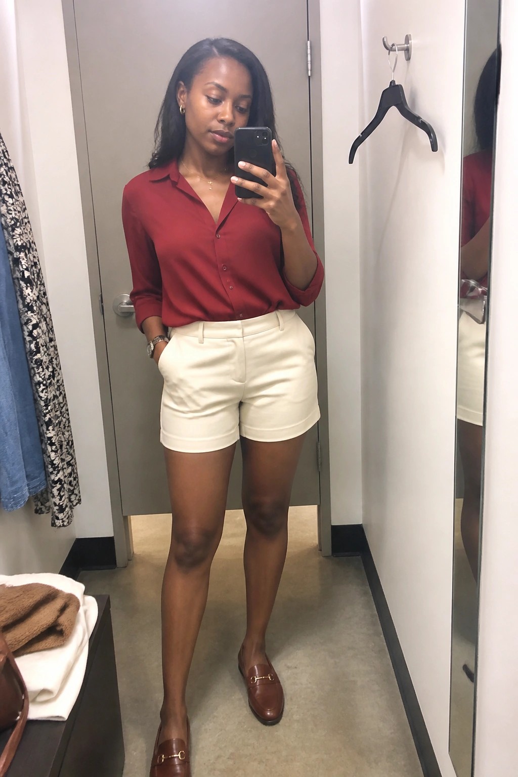 Woman in red shirt and white shorts taking mirror selfie in fitting room