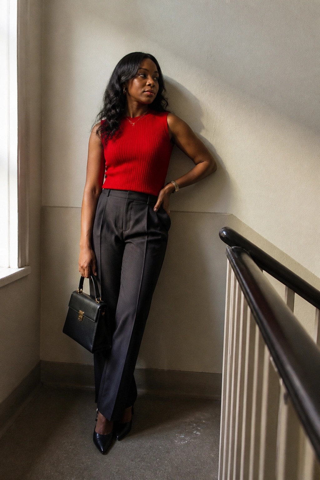 Woman in red sleeveless top and gray pants holds black handbag near stairs