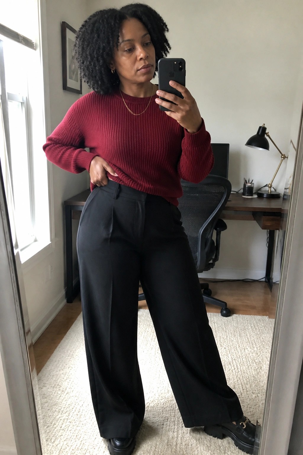 Woman in red sweater and black trousers taking mirror selfie in home office