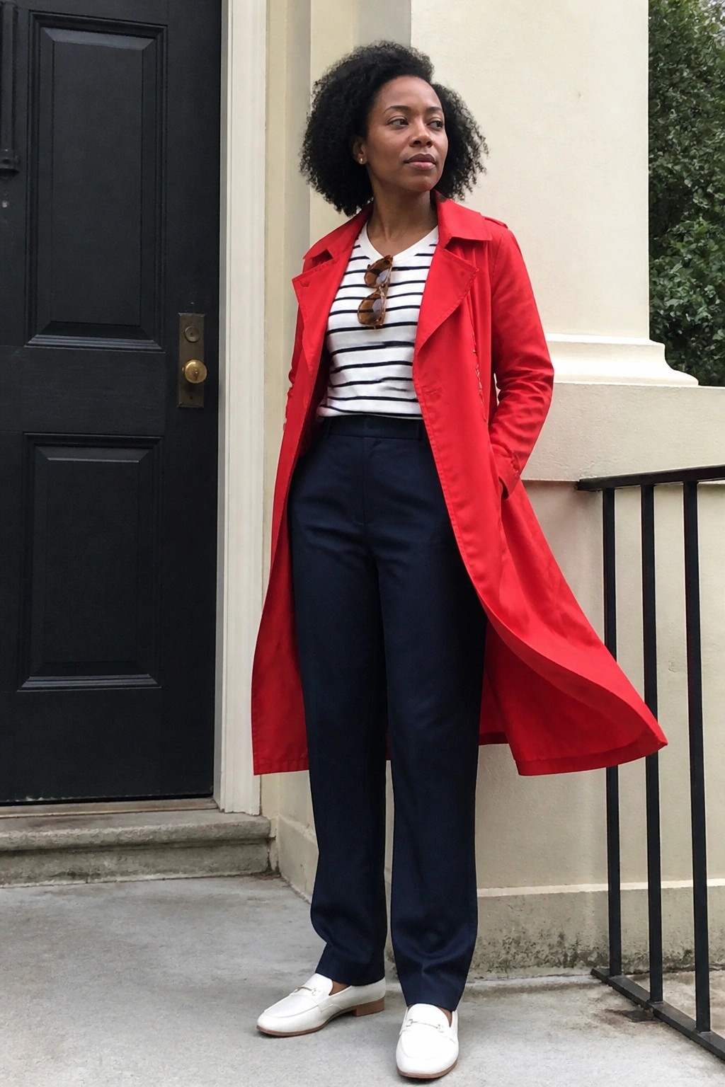 Woman in red trench coat over striped shirt with navy pants and white loafers outdoors
