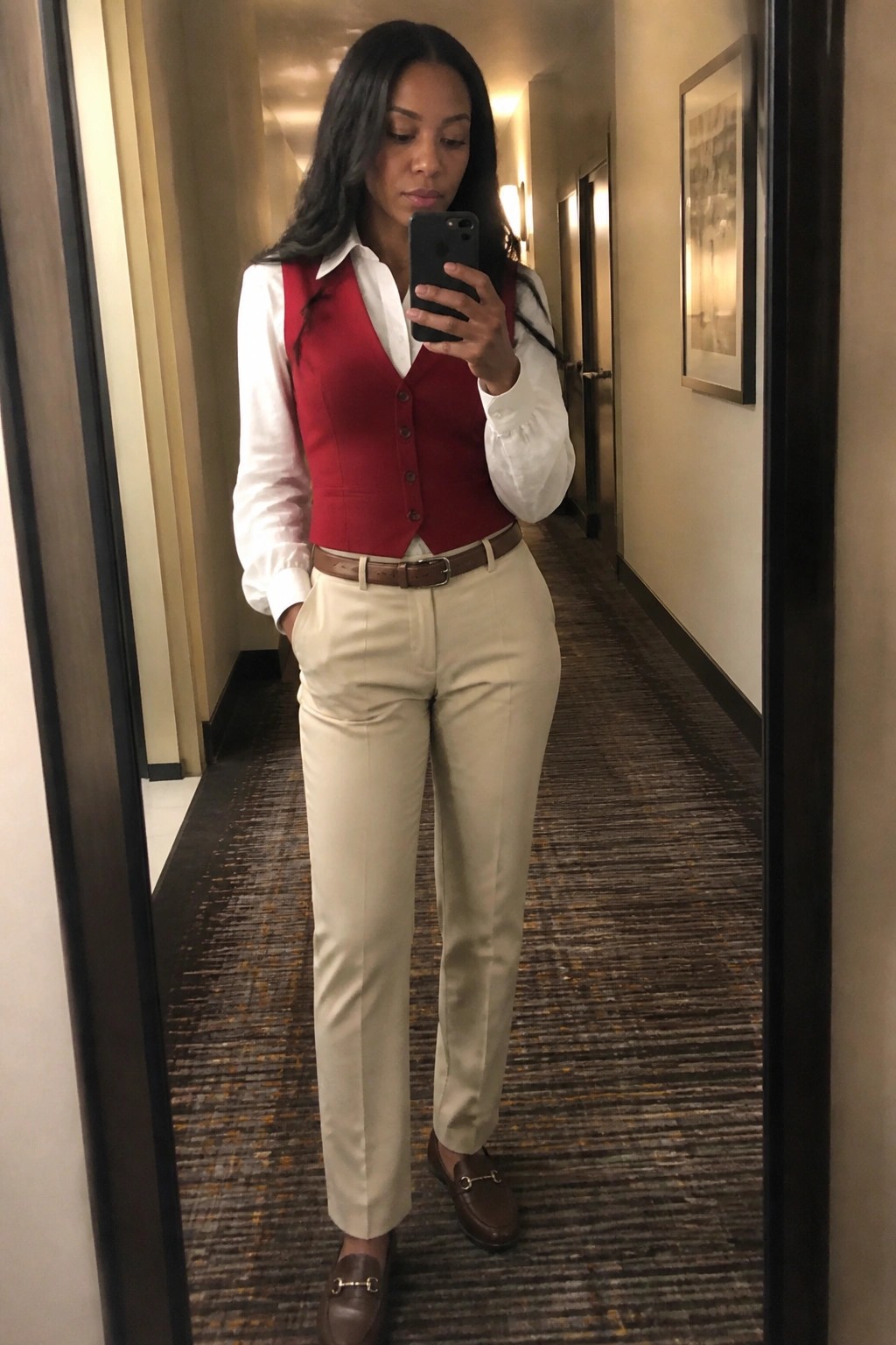 Woman in red vest, white shirt, and beige pants taking mirror selfie in hallway