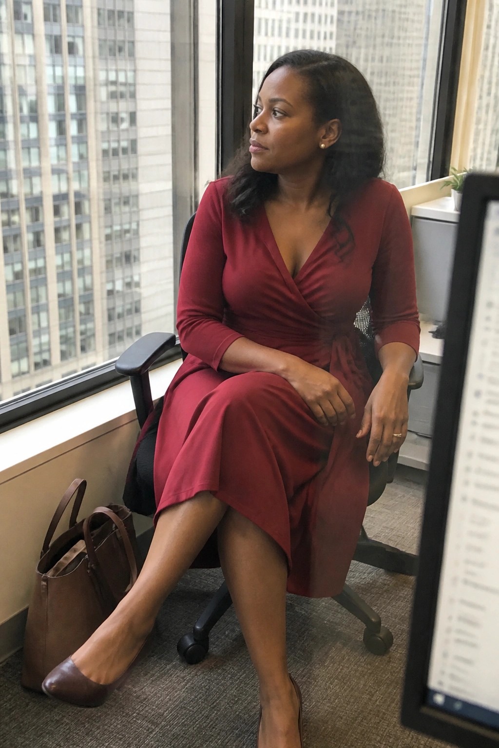 Woman in red wrap dress seated in office chair by window with brown handbag
