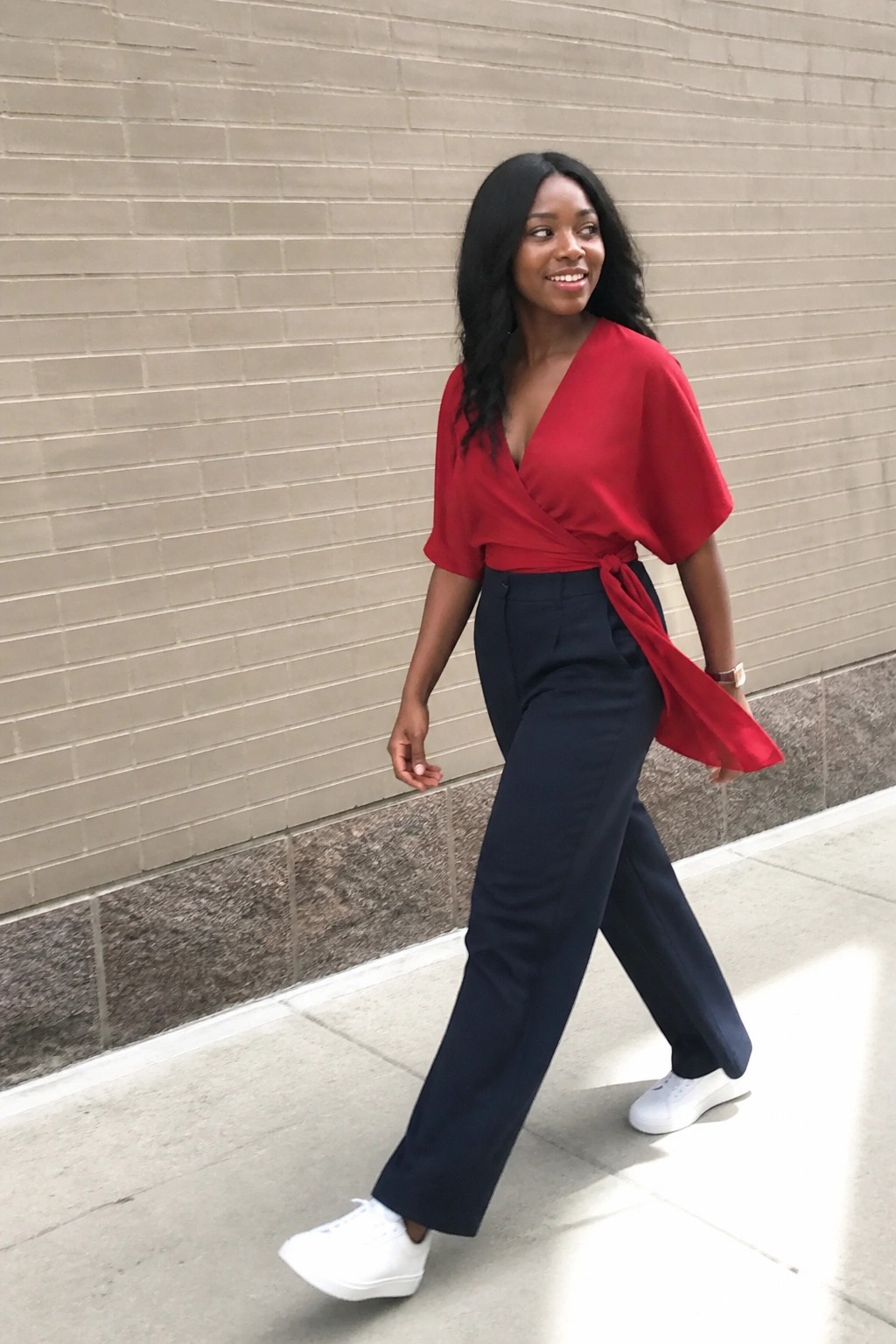 Woman wearing a red short sleeve wrap blouse tied at the waist with navy wide leg trousers and white sneakers standing in front of a brick wall.