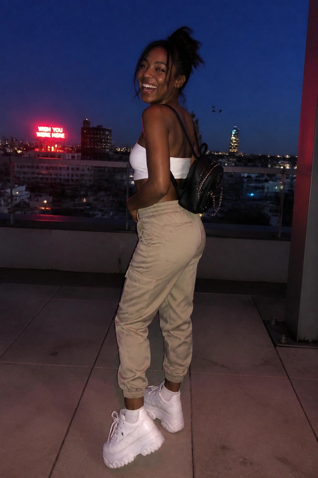 Woman in white crop top and beige cargo pants smiling on city rooftop at night