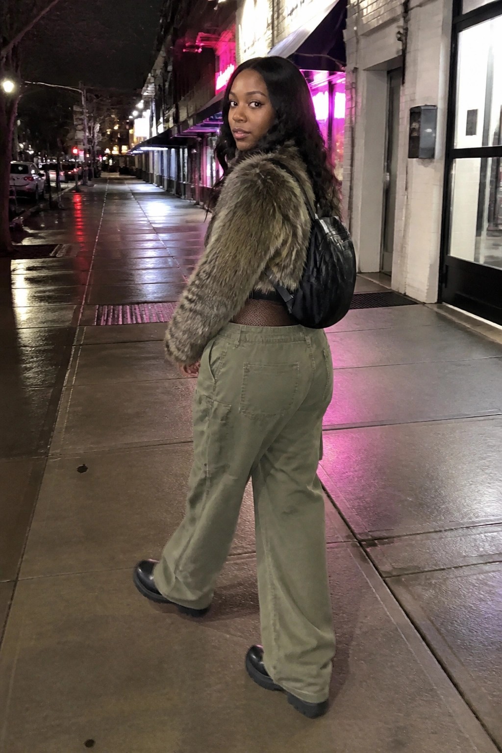 Woman in fur coat and green cargo pants with black bag on nighttime sidewalk