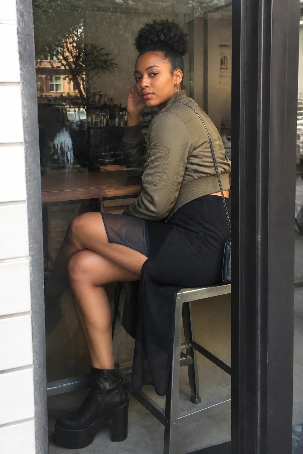 Woman in olive bomber jacket and sheer black skirt on metal stool by window