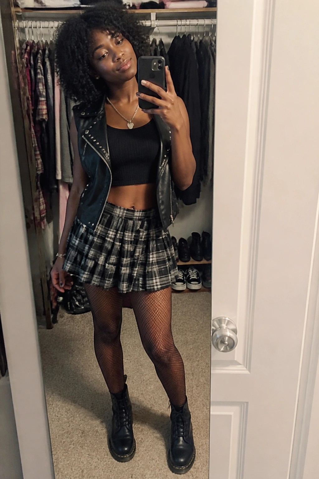 Woman in studded leather vest, plaid skirt, fishnets, and boots taking mirror selfie.
