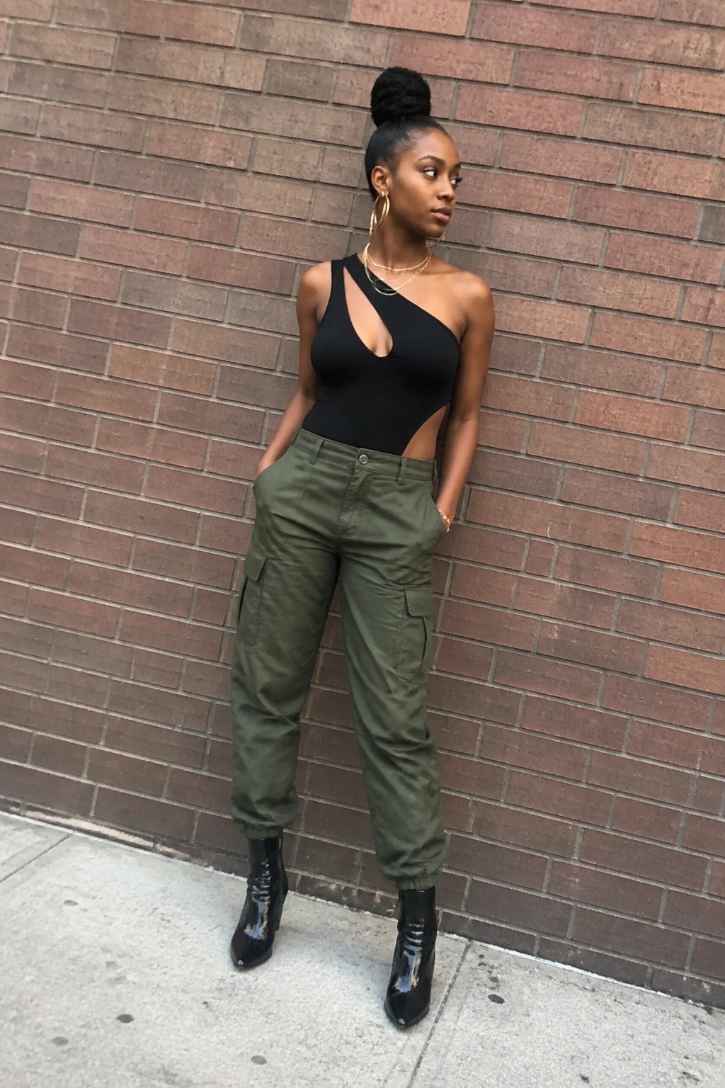 Woman in black one-shoulder cutout bodysuit tucked into olive cargo pants with black patent ankle boots and layered gold necklaces.