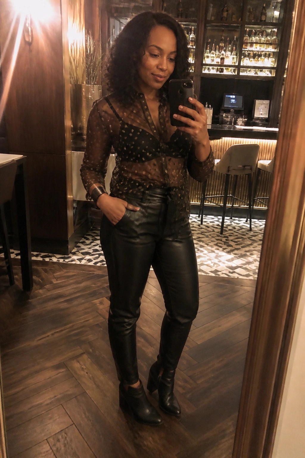 Woman in sheer polka-dot blouse and leather pants taking mirror selfie in bar