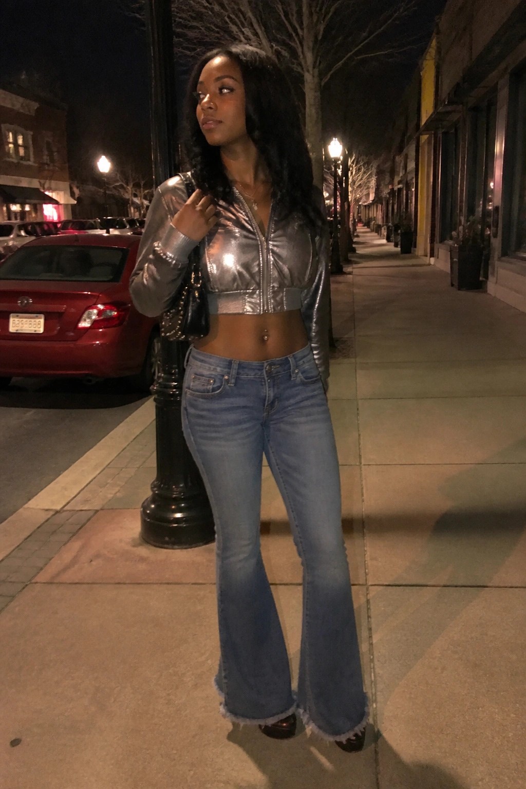Woman wearing a cropped silver metallic zip jacket with blue flared jeans and a chain strap bag on a city sidewalk at night.