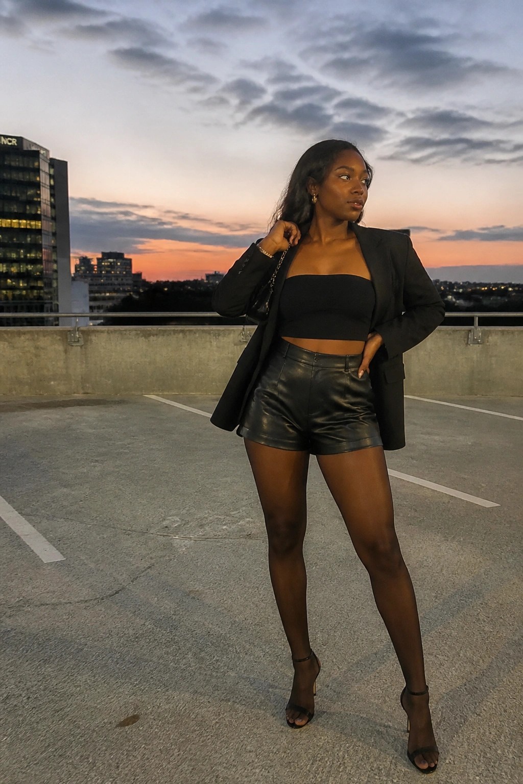 Stylish Black woman in black blazer and leather shorts posing on rooftop at sunset