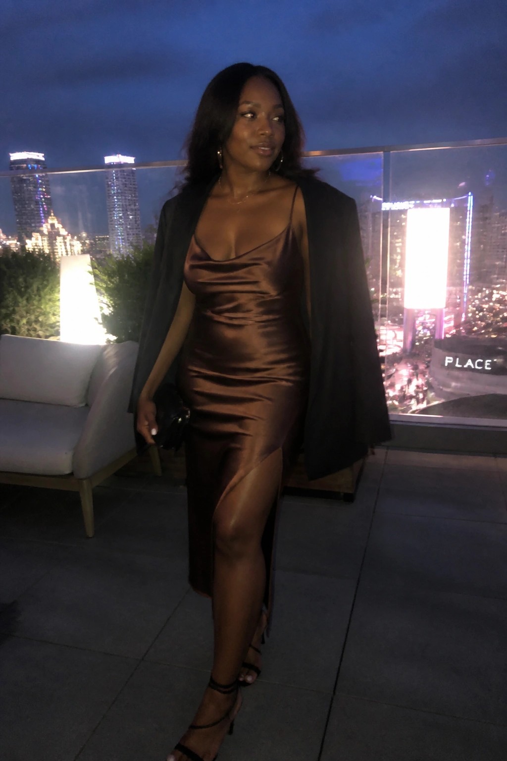 Woman in brown satin slip dress with black blazer on nighttime city balcony.