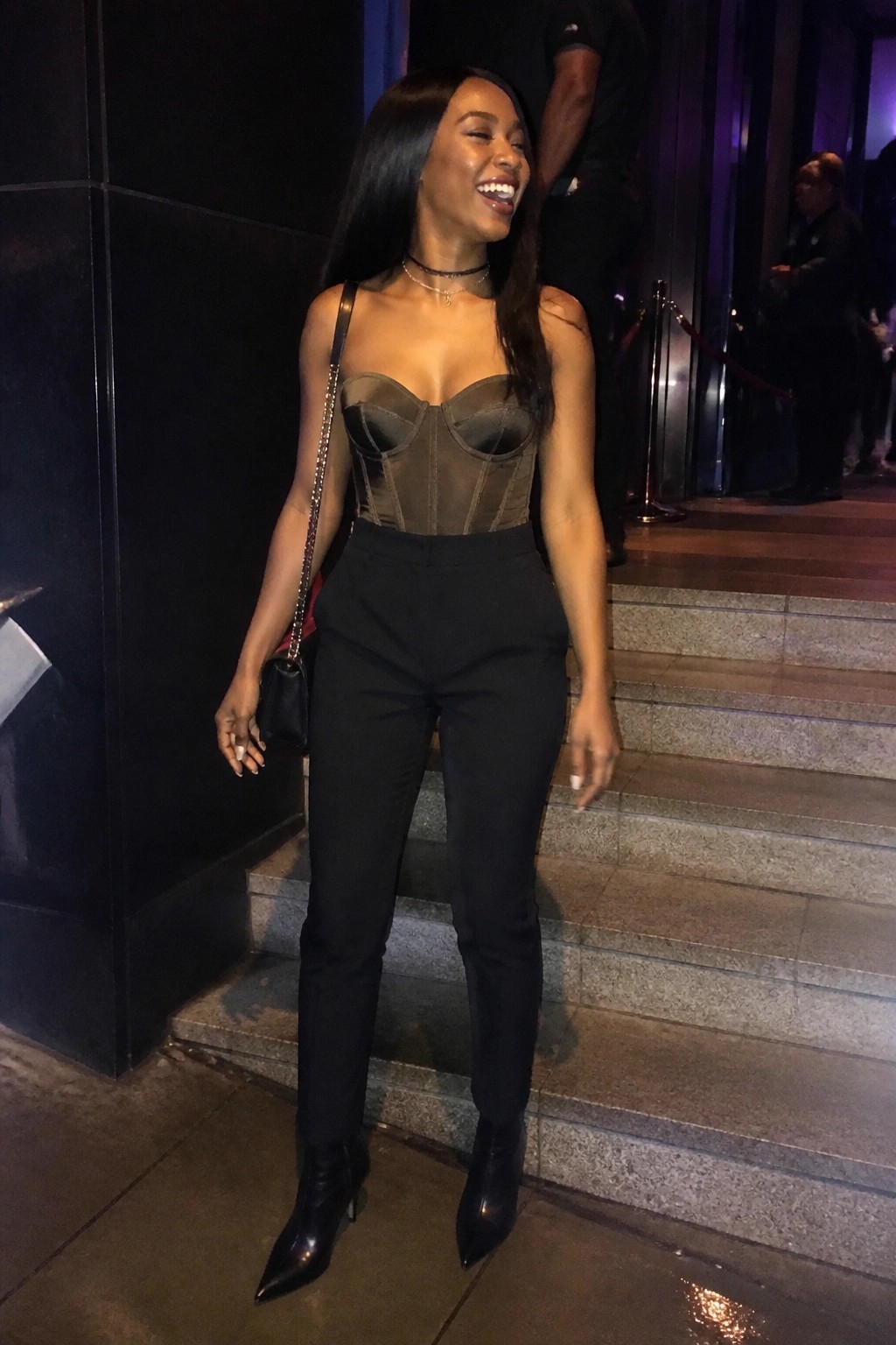 Woman wearing a black sheer corset top with structured cups, high-waisted slim black pants, pointed black ankle boots, and a small black chain strap handbag, standing on steps at night.