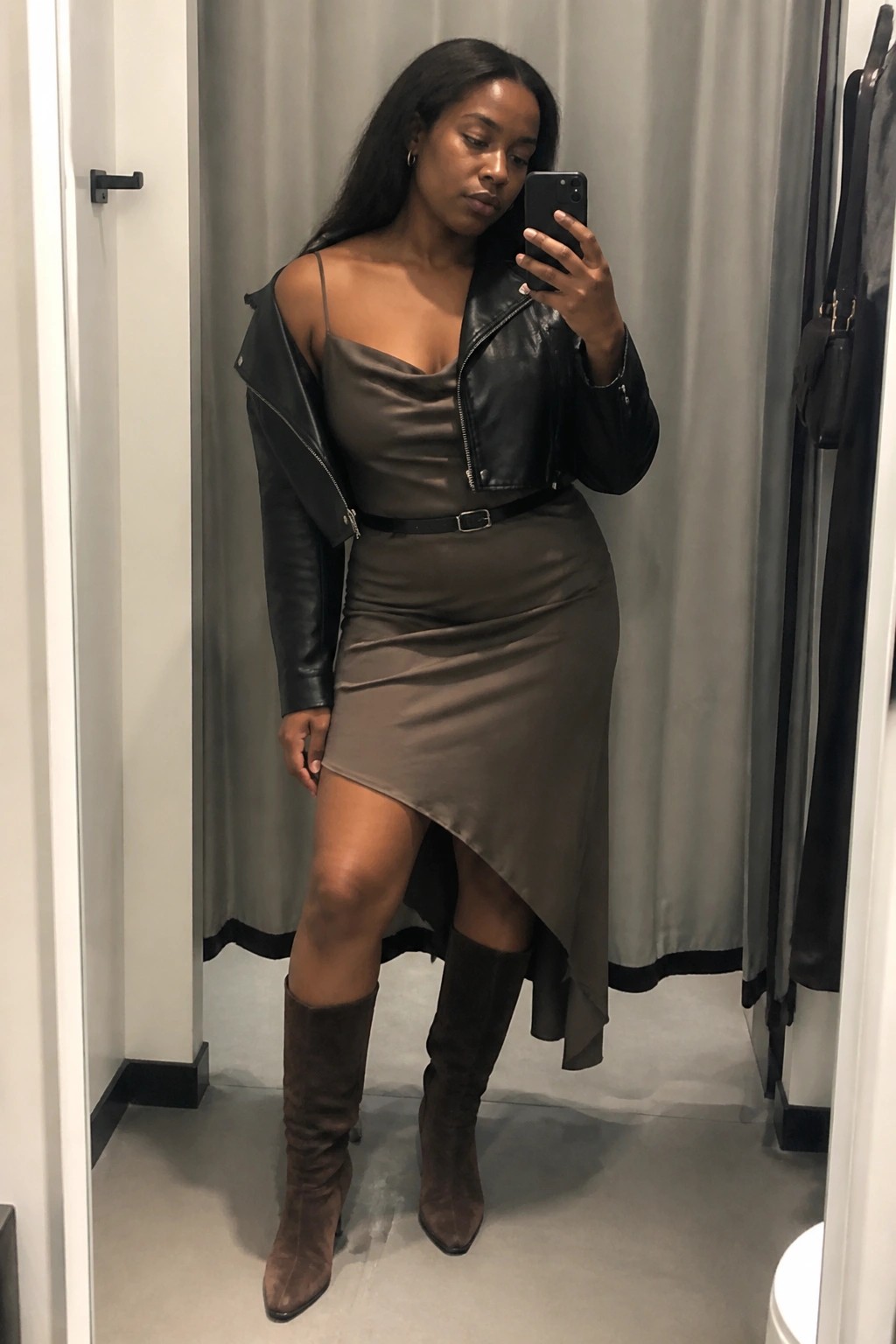 Woman in brown satin dress and black leather jacket taking mirror selfie in suede boots