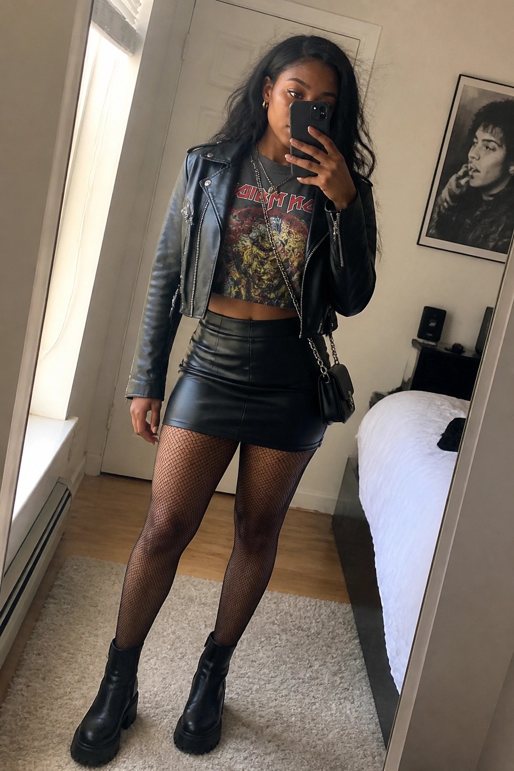 Woman in black leather jacket, mini skirt, fishnets, and boots taking bedroom mirror selfie