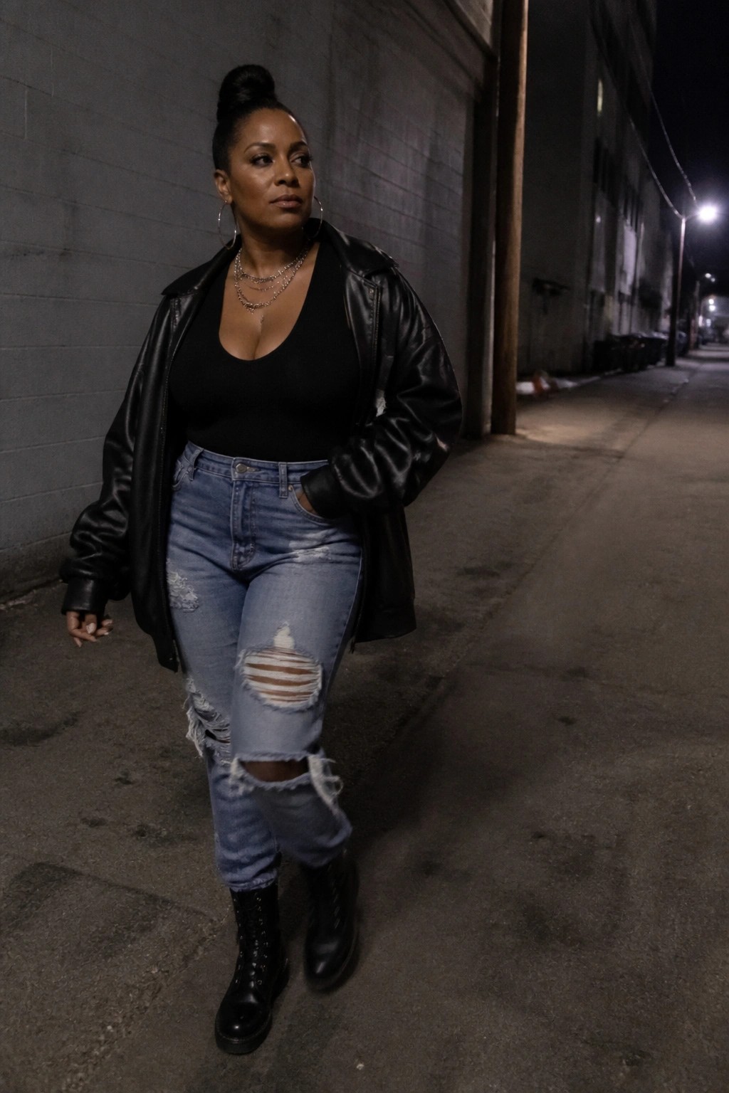 Woman in black leather jacket and ripped jeans stands in dark alley at night