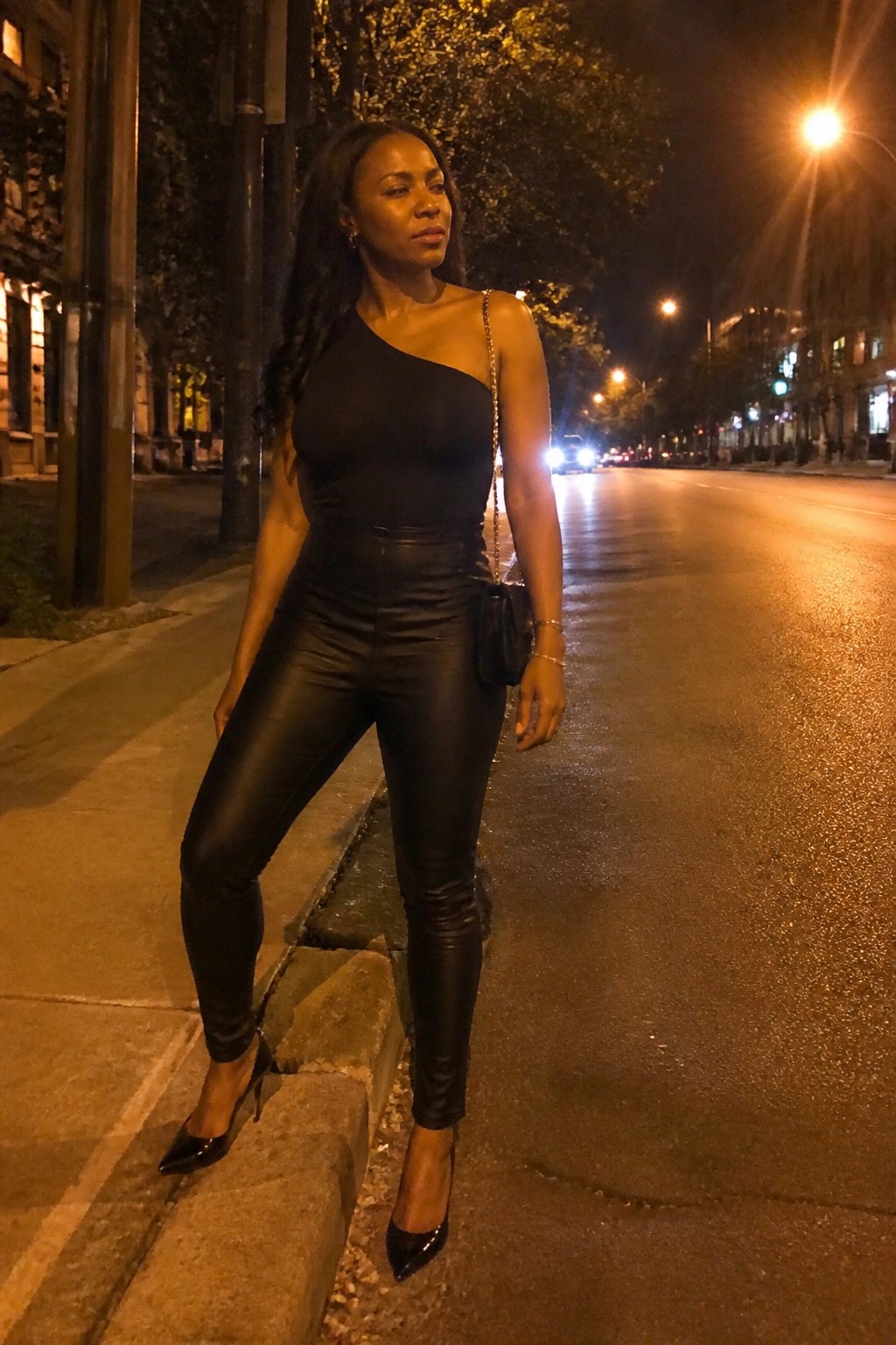 A woman stands on a city sidewalk at night wearing a black one-shoulder bodysuit tucked into black leather pants with pointed black heels and carrying a small black bag with a gold chain strap.