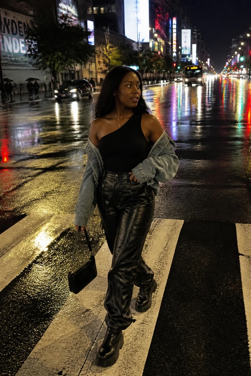 Woman in black one-shoulder top and leather pants crossing rainy city street at night