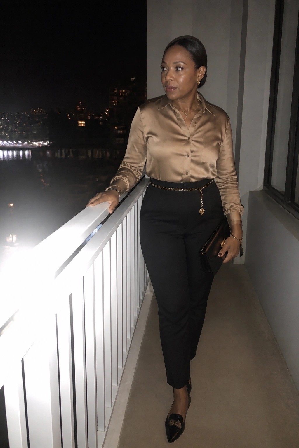 Woman in beige satin blouse and black pants with gold chain belt on night balcony