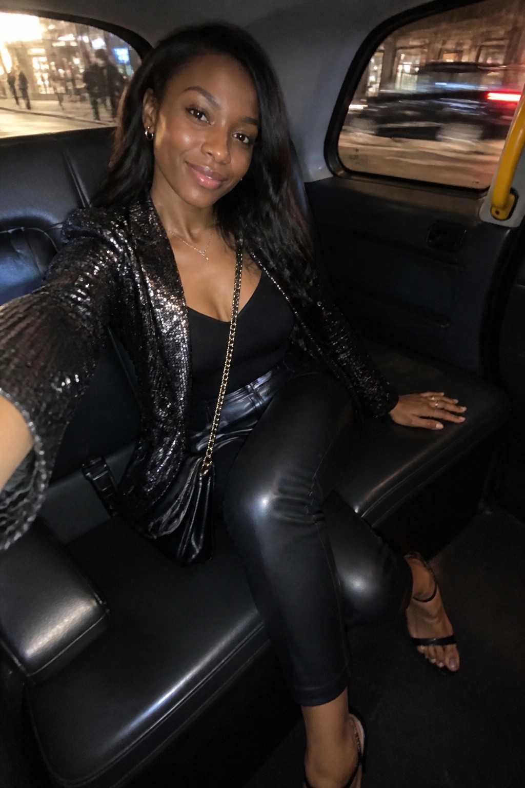 Black woman seated in a car wearing a black sequin jacket over a black tank top with black leather pants, gold chain purse, and black heeled sandals.