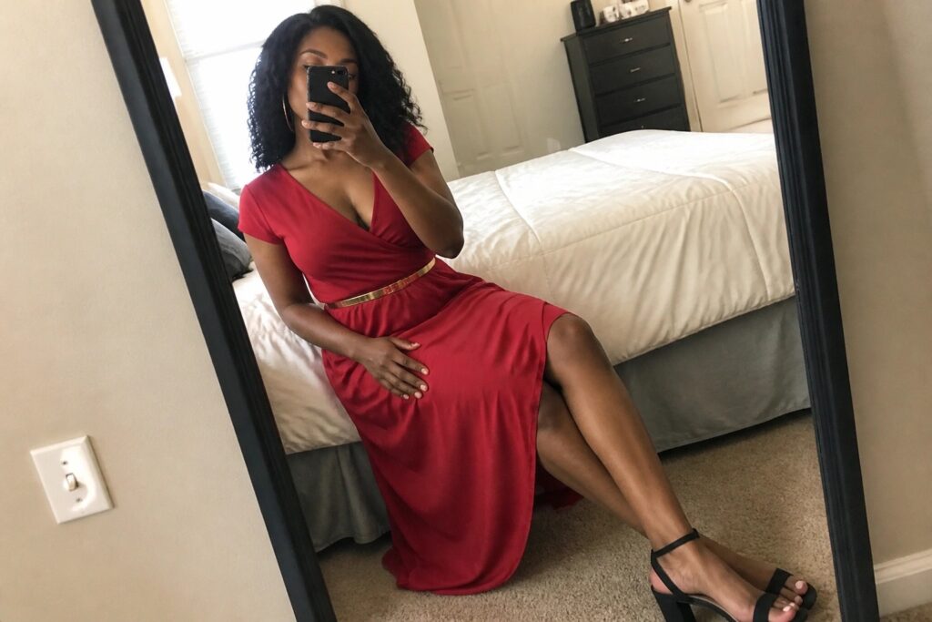 Woman in red wrap dress taking mirror selfie with phone in bedroom