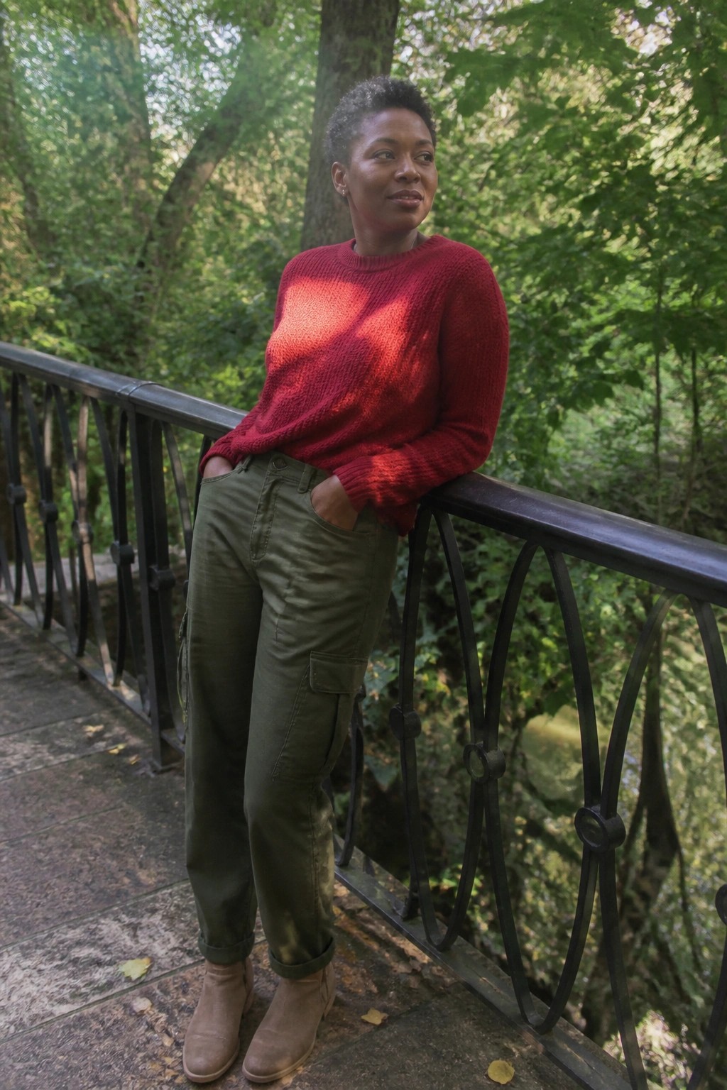 Woman in red sweater and green cargo pants leaning on railing in forest