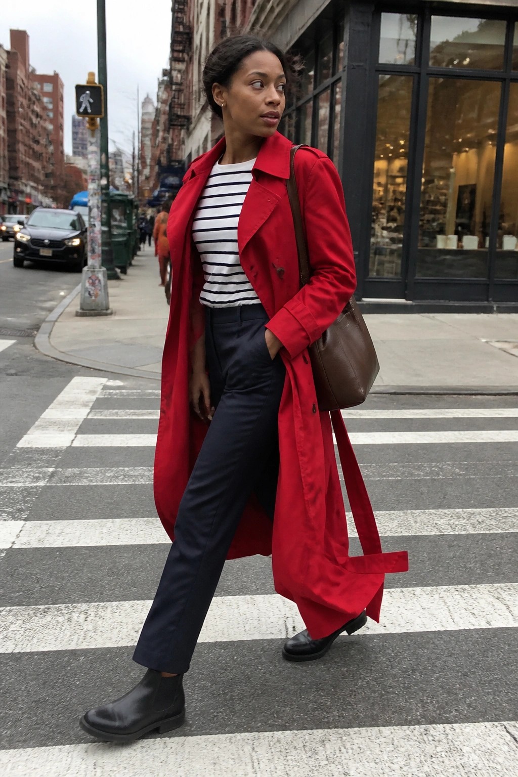 Woman in long red trench coat over striped top and navy trousers with black boots and brown leather bag on urban crosswalk