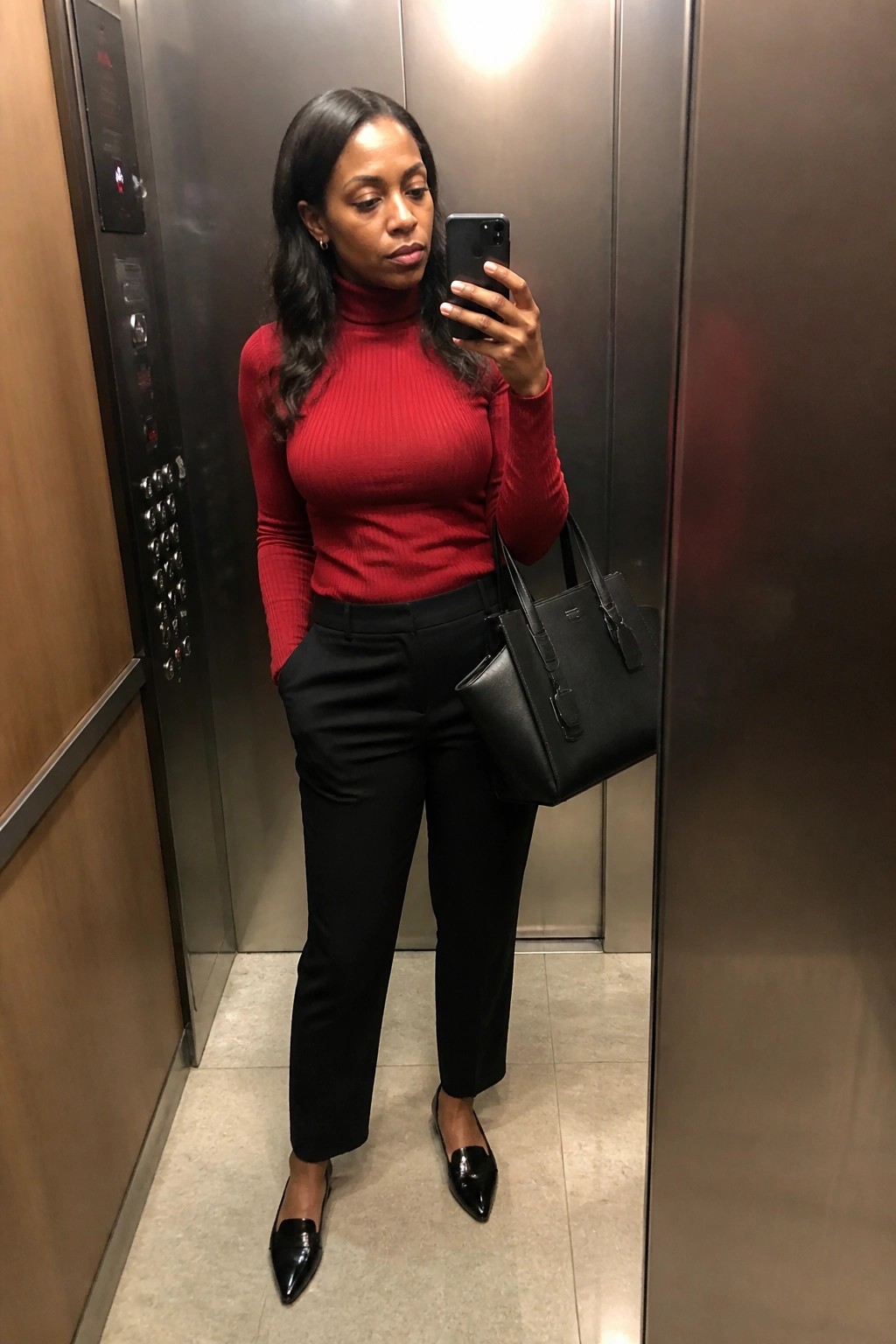 Woman in red turtleneck and black pants taking selfie in elevator with handbag
