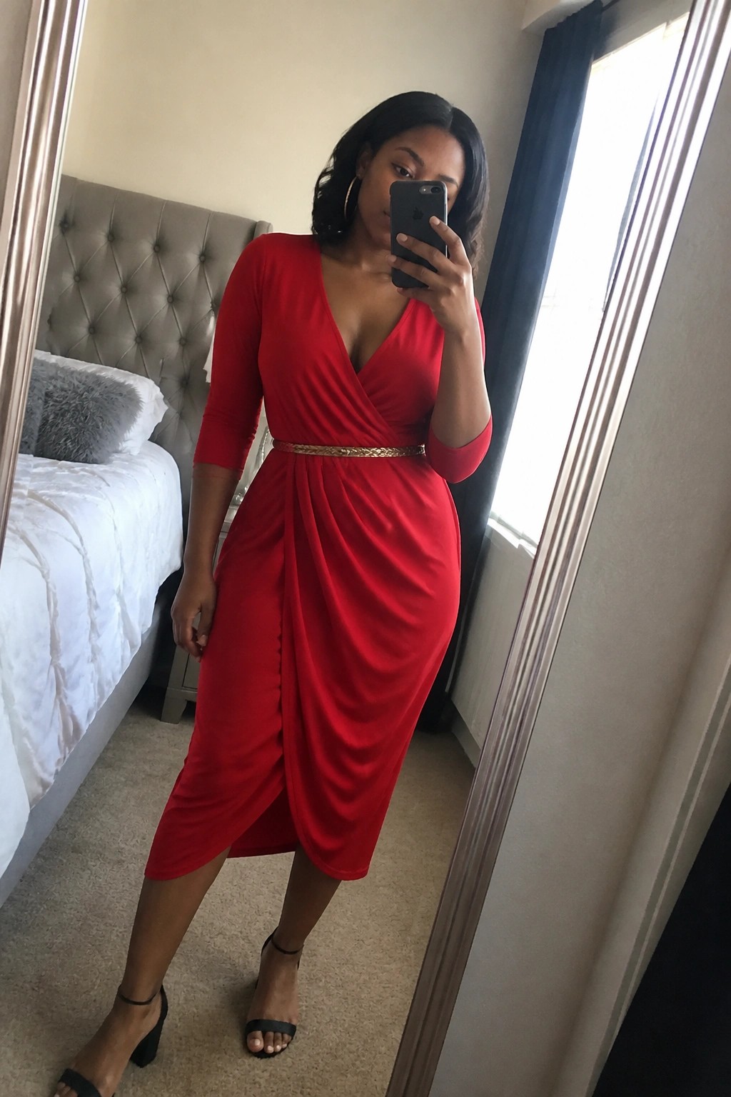 Woman in red wrap dress taking mirror selfie with phone in bedroom