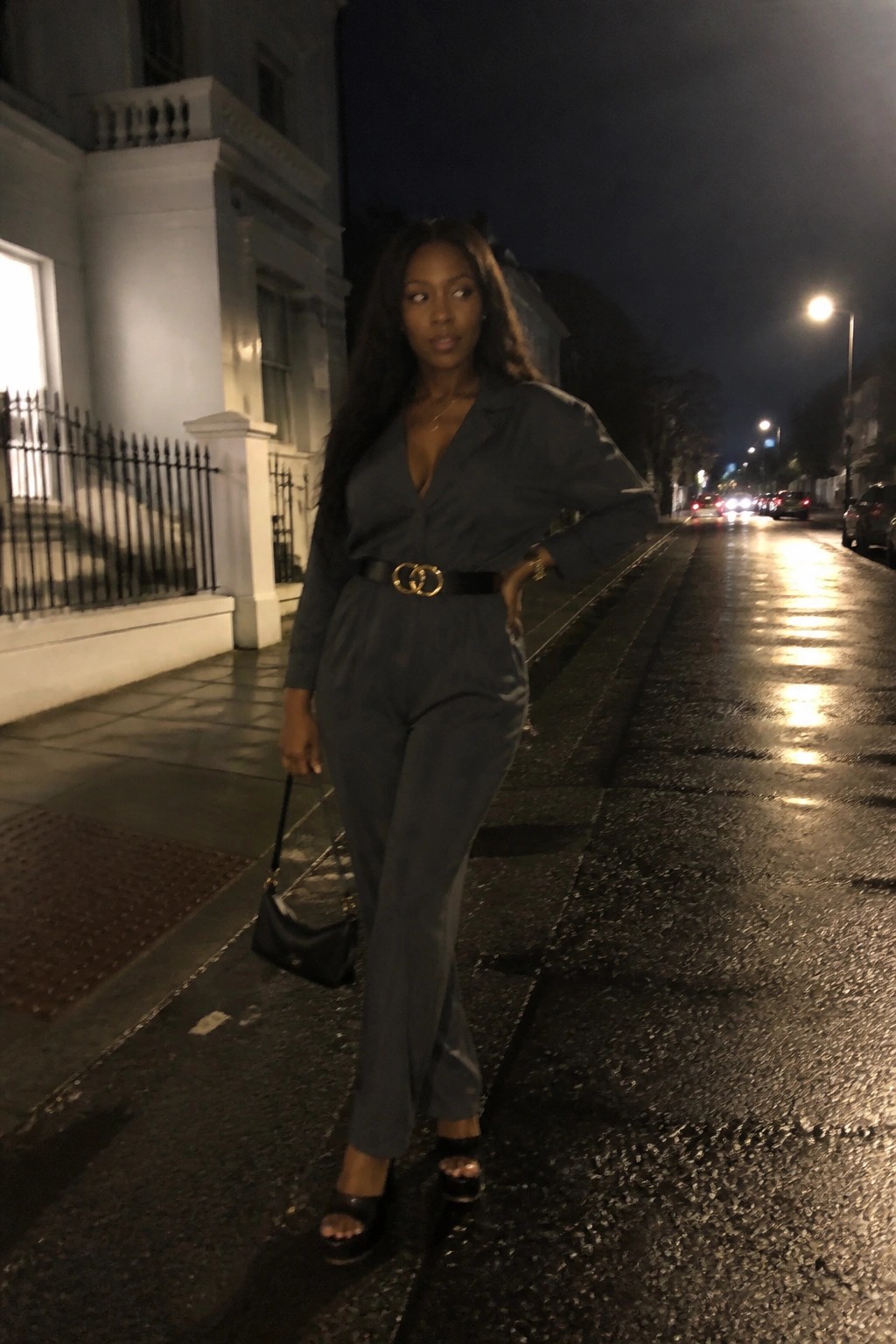 Woman in a charcoal gray belted jumpsuit with long sleeves deep V neck black platform heels and small black handbag standing on a wet city sidewalk at night.