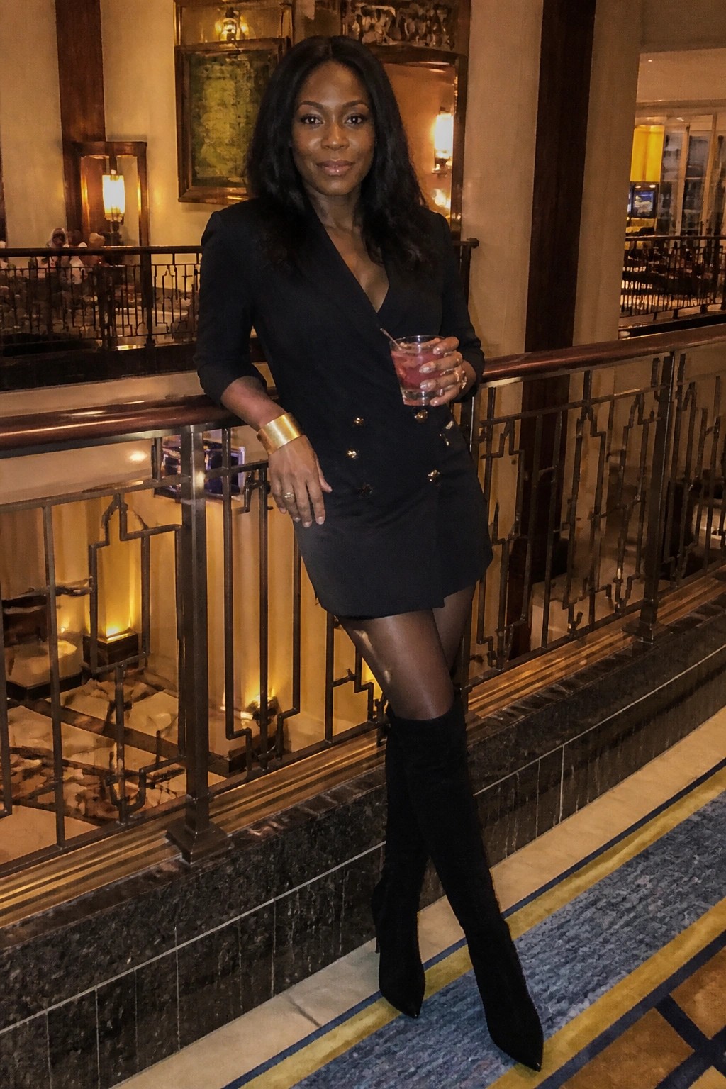 Woman in black blazer dress and boots holding cocktail on ornate balcony